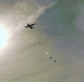 A large military transport aircraft, consistent with a C-130 Hercules silhouette, is photographed from below against a bright, hazy sky with the sun visible at left. The aircraft appears to be conducting an airdrop operation, with multiple parachutes or cargo bundles visible descending below the aircraft. The image is backlit and slightly overexposed due to the sun's proximity in the frame.