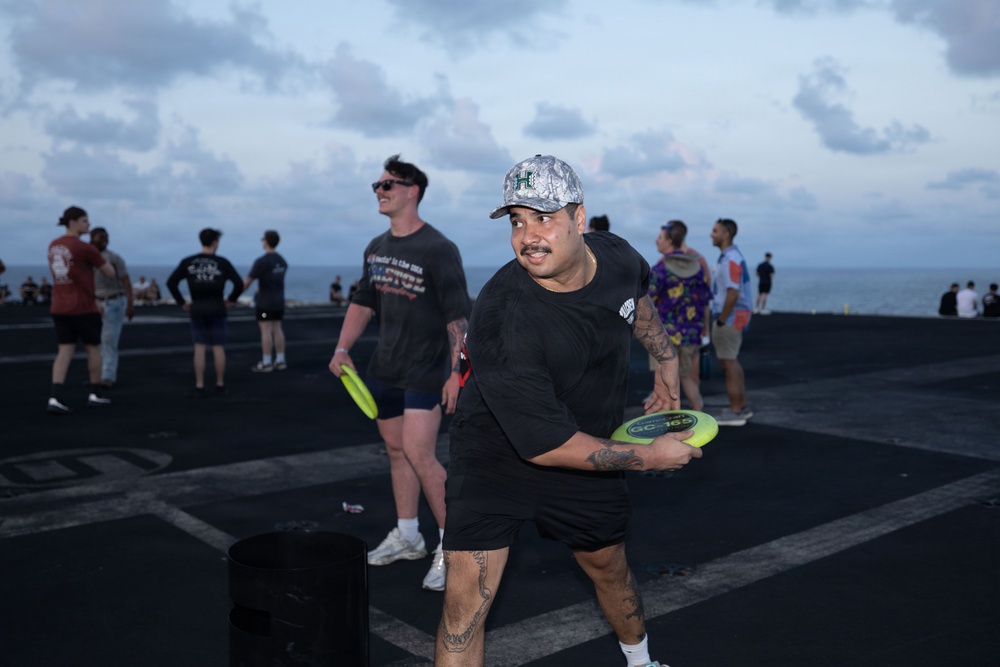 A group of people are playing frisbee on what appears to be the flight deck of a naval vessel at dusk, with the ocean visible in the background. The foreground shows a man in a black outfit and camo cap holding a yellow-green frisbee in a throwing stance. Multiple other individuals are visible in the background engaged in similar recreational activities.