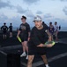A group of people are playing frisbee on what appears to be the flight deck of a naval vessel at dusk, with the ocean visible in the background. The foreground shows a man in a black outfit and camo cap holding a yellow-green frisbee in a throwing stance. Multiple other individuals are visible in the background engaged in similar recreational activities.