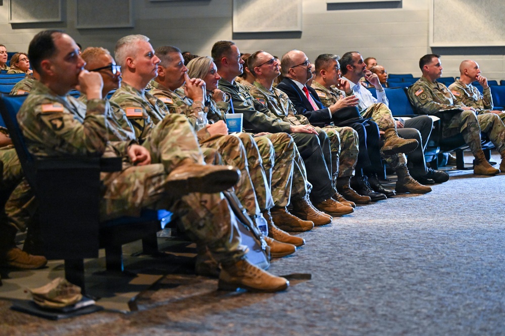 A group of U.S. Army military personnel and civilian attendees are seated in an auditorium or conference room, appearing to listen to a presentation or briefing. The majority are wearing Army Combat Uniforms (ACU) in OCP (Operational Camouflage Pattern), with a few civilians in business attire visible among them. The audience appears attentive, with several individuals in contemplative or listening postures.