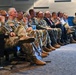 A group of U.S. Army military personnel and civilian attendees are seated in an auditorium or conference room, appearing to listen to a presentation or briefing. The majority are wearing Army Combat Uniforms (ACU) in OCP (Operational Camouflage Pattern), with a few civilians in business attire visible among them. The audience appears attentive, with several individuals in contemplative or listening postures.