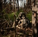A soldier in full combat gear is positioned in a prone or low-crouching stance within a wooded forest environment, aiming a rifle equipped with an optical scope. A second soldier is partially visible in the background to the left. The image appears to depict a military field exercise or tactical operation in a forested setting.