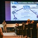 A military service member in camouflage uniform is presenting a briefing slide titled 'UC26 FTX Medical Thread Role 1 to Role 4' to an audience seated in a theater-style room. The presentation outlines a medical evacuation and patient movement exercise involving multiple military installations including Fort Bragg, Fort Campbell, Scott AFB IL, and Alpena MI. The slide depicts a logistics flow diagram showing medical roles and transportation methods across CONUS and OCONUS locations.