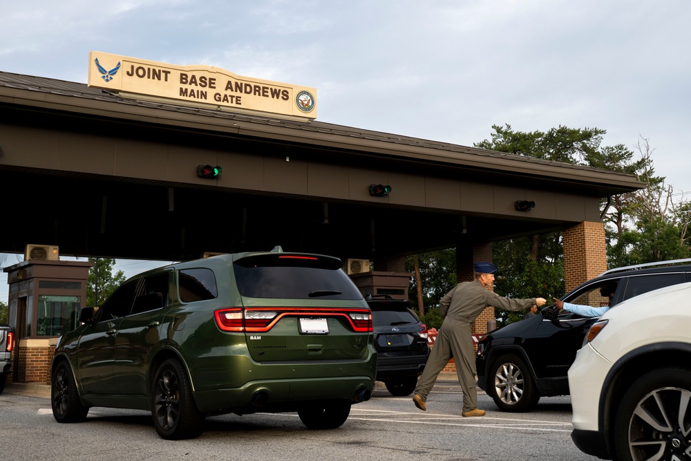 The image shows the Main Gate entrance of Joint Base Andrews, a U.S. military installation. A person in a flight suit or military uniform is leaning toward a vehicle window, likely conducting an access control check. Multiple vehicles are queued at the gate checkpoint under an overhead sign displaying the base name and military branch insignia.
