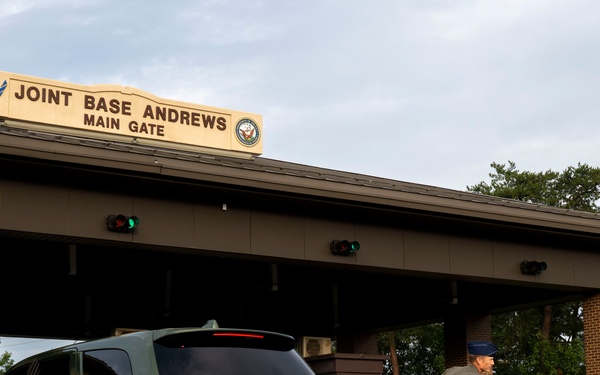 The image shows the Main Gate entrance of Joint Base Andrews, a U.S. military installation. A person in a flight suit or military uniform is leaning toward a vehicle window, likely conducting an access control check. Multiple vehicles are queued at the gate checkpoint under an overhead sign displaying the base name and military branch insignia.