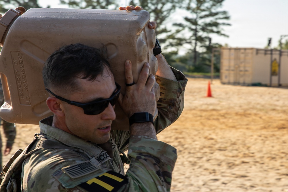 A U.S. Army soldier in MultiCam/OCP uniform is carrying a large tan jerry can or water container marked 'Property of U.S. Government' on his shoulder during what appears to be a physical training or field exercise. The setting is an outdoor sandy training area with shipping containers and orange traffic cones visible in the background. The soldier is wearing sunglasses and a fitness tracker on his wrist.