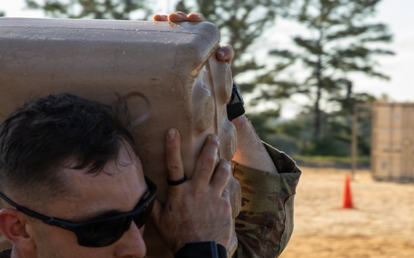 A U.S. Army soldier in MultiCam/OCP uniform is carrying a large tan jerry can or water container marked 'Property of U.S. Government' on his shoulder during what appears to be a physical training or field exercise. The setting is an outdoor sandy training area with shipping containers and orange traffic cones visible in the background. The soldier is wearing sunglasses and a fitness tracker on his wrist.