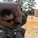 A U.S. Army soldier in MultiCam/OCP uniform is carrying a large tan jerry can or water container marked 'Property of U.S. Government' on his shoulder during what appears to be a physical training or field exercise. The setting is an outdoor sandy training area with shipping containers and orange traffic cones visible in the background. The soldier is wearing sunglasses and a fitness tracker on his wrist.