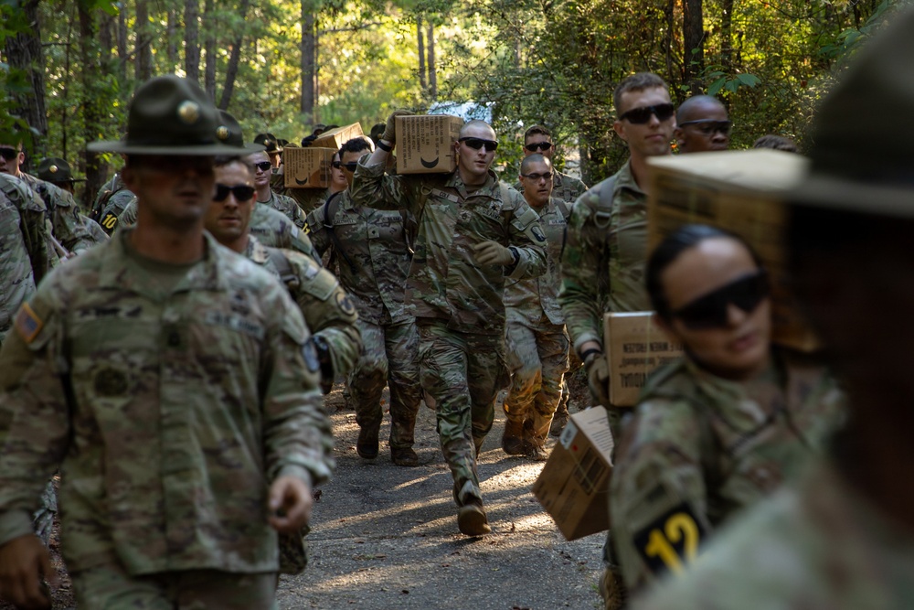A group of U.S. Army soldiers in camouflage uniforms are marching along a wooded trail while carrying cardboard boxes, likely MRE (Meal Ready-to-Eat) rations or supply crates. Drill sergeants wearing distinctive campaign (Smokey Bear) hats are visible supervising the formation. This appears to be a training exercise or field logistics activity at a military installation.