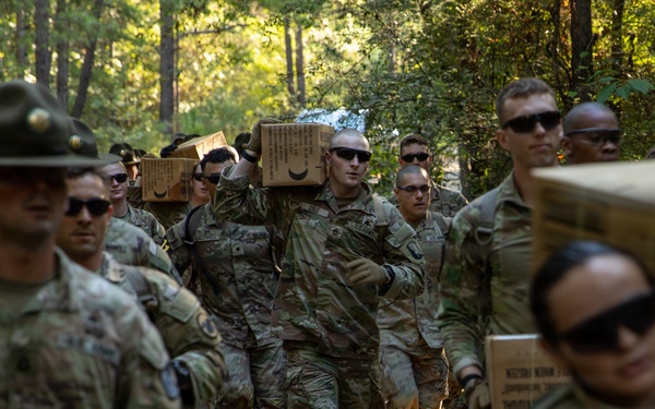 A group of U.S. Army soldiers in camouflage uniforms are marching along a wooded trail while carrying cardboard boxes, likely MRE (Meal Ready-to-Eat) rations or supply crates. Drill sergeants wearing distinctive campaign (Smokey Bear) hats are visible supervising the formation. This appears to be a training exercise or field logistics activity at a military installation.