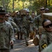 A group of U.S. Army soldiers in camouflage uniforms are marching along a wooded trail while carrying cardboard boxes, likely MRE (Meal Ready-to-Eat) rations or supply crates. Drill sergeants wearing distinctive campaign (Smokey Bear) hats are visible supervising the formation. This appears to be a training exercise or field logistics activity at a military installation.