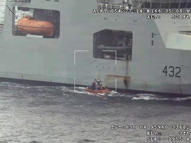 An aerial surveillance image captured from an aircraft showing a large grey naval vessel numbered '432' at sea. A small orange rigid inflatable boat (RIB) with personnel aboard is positioned alongside the larger ship near an open bay or hatch on the hull. The image contains telemetry overlay data indicating aircraft position, altitude (521ft), and target coordinates.