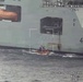 An aerial surveillance image captured from an aircraft showing a large grey naval vessel numbered '432' at sea. A small orange rigid inflatable boat (RIB) with personnel aboard is positioned alongside the larger ship near an open bay or hatch on the hull. The image contains telemetry overlay data indicating aircraft position, altitude (521ft), and target coordinates.