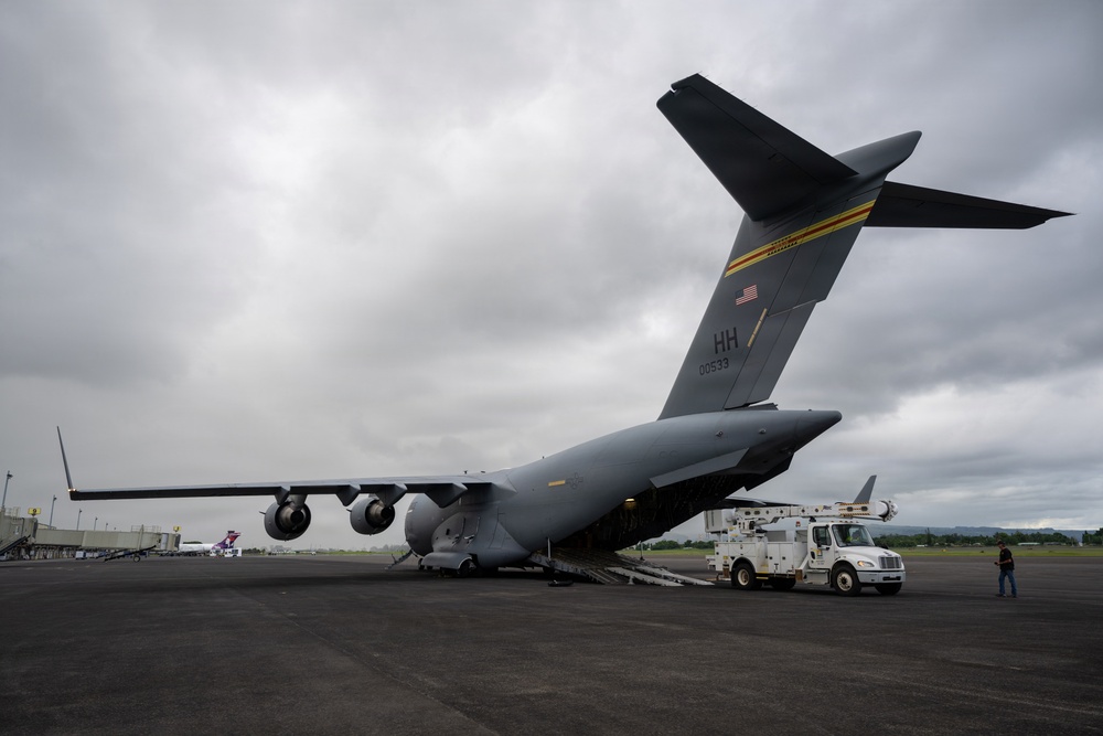 A U.S. Air Force C-17 Globemaster III military transport aircraft with tail code 'HH' and serial number 00533 is parked on a tarmac with its rear cargo ramp lowered. A utility/maintenance truck is positioned at the open cargo ramp, suggesting active loading or unloading operations. The scene appears to be at a civilian or joint-use airport, with overcast skies and what appears to be a Hawaiian Airlines aircraft visible in the background.