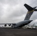 A U.S. Air Force C-17 Globemaster III military transport aircraft with tail code 'HH' and serial number 00533 is parked on a tarmac with its rear cargo ramp lowered. A utility/maintenance truck is positioned at the open cargo ramp, suggesting active loading or unloading operations. The scene appears to be at a civilian or joint-use airport, with overcast skies and what appears to be a Hawaiian Airlines aircraft visible in the background.