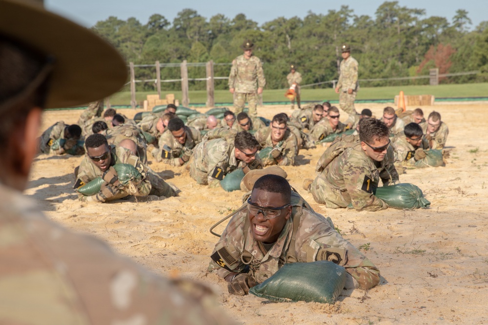 A large group of military trainees in camouflage uniforms are performing a low-crawl exercise on a sandy training ground while carrying green sandbags. Drill instructors or cadre members are standing and observing the exercise from the background. The setting appears to be a military training facility, likely a basic training or initial entry training environment.