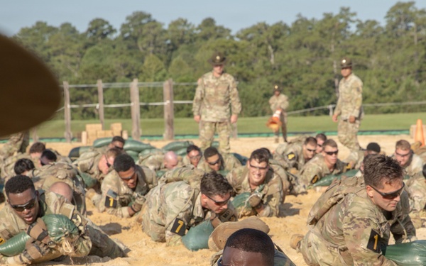 A large group of military trainees in camouflage uniforms are performing a low-crawl exercise on a sandy training ground while carrying green sandbags. Drill instructors or cadre members are standing and observing the exercise from the background. The setting appears to be a military training facility, likely a basic training or initial entry training environment.