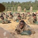 A large group of military trainees in camouflage uniforms are performing a low-crawl exercise on a sandy training ground while carrying green sandbags. Drill instructors or cadre members are standing and observing the exercise from the background. The setting appears to be a military training facility, likely a basic training or initial entry training environment.