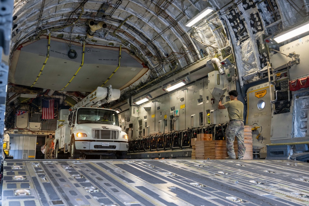 The interior of a large military cargo aircraft, likely a C-17 Globemaster III, is shown being loaded with a white utility/bucket truck and palletized cargo. Two personnel in military uniforms are visible inside the aircraft. American and British flags are displayed near the front of the cargo hold, suggesting a joint operation or humanitarian mission.