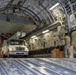 The interior of a large military cargo aircraft, likely a C-17 Globemaster III, is shown being loaded with a white utility/bucket truck and palletized cargo. Two personnel in military uniforms are visible inside the aircraft. American and British flags are displayed near the front of the cargo hold, suggesting a joint operation or humanitarian mission.