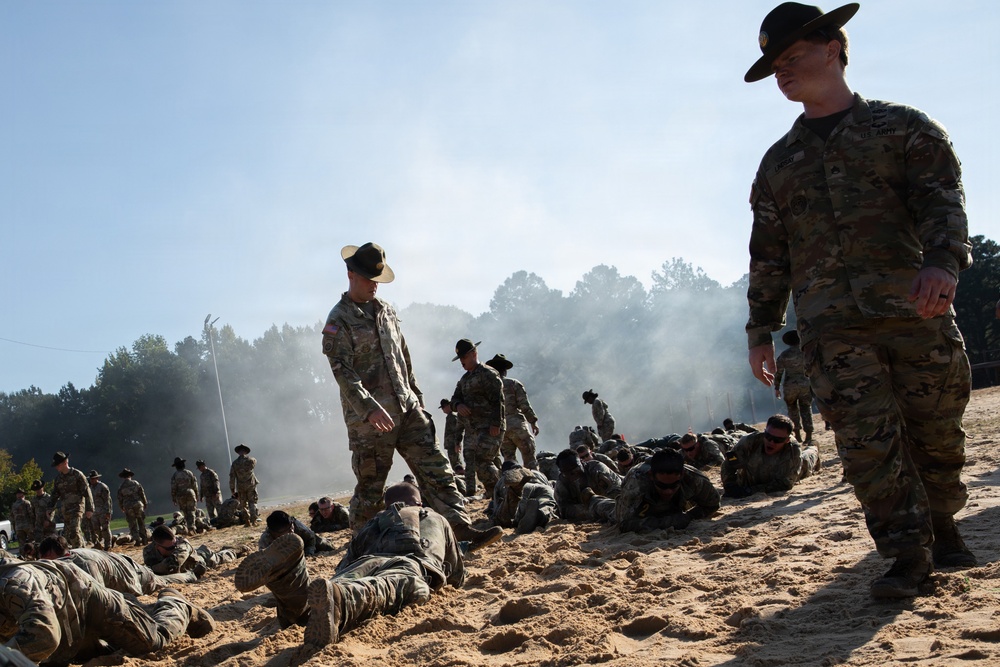The image depicts a U.S. Army basic training or physical fitness exercise on a sandy terrain. Drill sergeants wearing distinctive campaign (Smokey Bear) hats are overseeing recruits who are low-crawling or performing ground exercises in the sand. Smoke is visible in the background, suggesting a training exercise scenario.