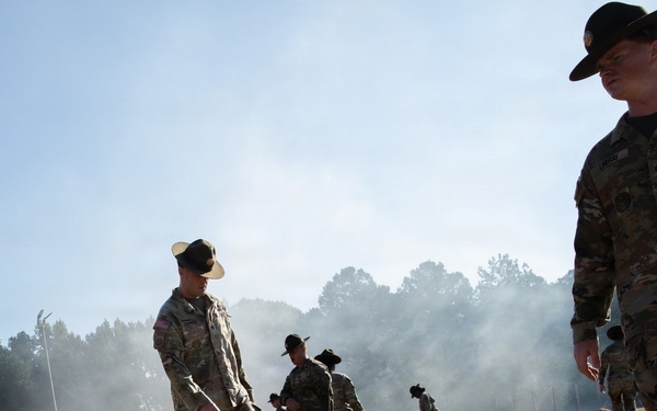 The image depicts a U.S. Army basic training or physical fitness exercise on a sandy terrain. Drill sergeants wearing distinctive campaign (Smokey Bear) hats are overseeing recruits who are low-crawling or performing ground exercises in the sand. Smoke is visible in the background, suggesting a training exercise scenario.