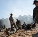 The image depicts a U.S. Army basic training or physical fitness exercise on a sandy terrain. Drill sergeants wearing distinctive campaign (Smokey Bear) hats are overseeing recruits who are low-crawling or performing ground exercises in the sand. Smoke is visible in the background, suggesting a training exercise scenario.