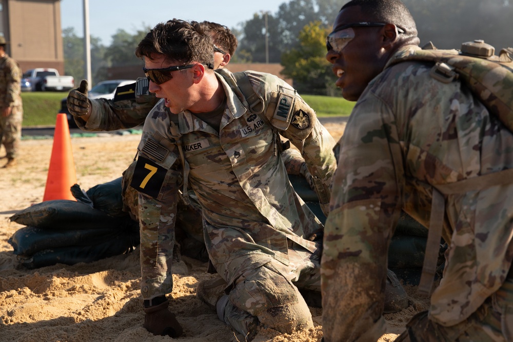 U.S. Army Military Police soldiers are engaged in a physical training or competition event on a sandy surface. The central soldier, identified by the name tape 'WALKER' and wearing number '7' on an armband, appears to be performing a strenuous physical task while another soldier observes or participates nearby. The setting appears to be a military installation with buildings and vehicles visible in the background.