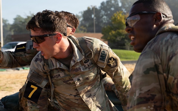 U.S. Army Military Police soldiers are engaged in a physical training or competition event on a sandy surface. The central soldier, identified by the name tape 'WALKER' and wearing number '7' on an armband, appears to be performing a strenuous physical task while another soldier observes or participates nearby. The setting appears to be a military installation with buildings and vehicles visible in the background.