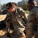 U.S. Army Military Police soldiers are engaged in a physical training or competition event on a sandy surface. The central soldier, identified by the name tape 'WALKER' and wearing number '7' on an armband, appears to be performing a strenuous physical task while another soldier observes or participates nearby. The setting appears to be a military installation with buildings and vehicles visible in the background.