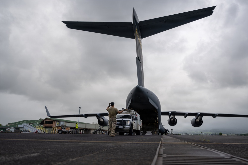A military cargo aircraft, consistent with a C-17 Globemaster III, is photographed from a low rear angle on a tarmac under overcast skies. A uniformed personnel member stands near the open rear cargo ramp while a white pickup truck is being loaded or unloaded from the aircraft. Airport infrastructure and green buildings are visible in the background.