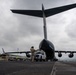 A military cargo aircraft, consistent with a C-17 Globemaster III, is photographed from a low rear angle on a tarmac under overcast skies. A uniformed personnel member stands near the open rear cargo ramp while a white pickup truck is being loaded or unloaded from the aircraft. Airport infrastructure and green buildings are visible in the background.
