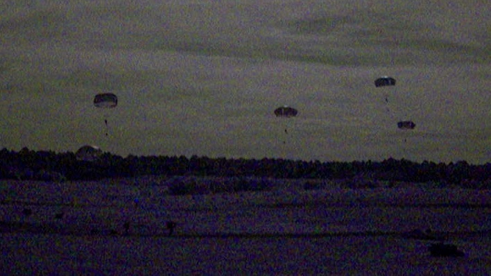 A low-light, grainy image captured at dusk or dawn showing multiple parachutes descending over a flat, open landscape. At least four parachutes are visible at varying altitudes, suggesting a military or training airborne operation. The poor lighting and image noise significantly reduce detail visibility.