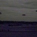 A low-light, grainy image captured at dusk or dawn showing multiple parachutes descending over a flat, open landscape. At least four parachutes are visible at varying altitudes, suggesting a military or training airborne operation. The poor lighting and image noise significantly reduce detail visibility.