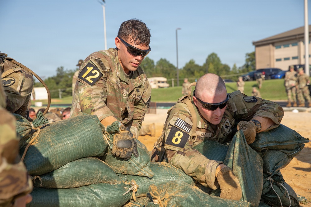 Two U.S. Army soldiers wearing OCP (Operational Camouflage Pattern) uniforms and numbered competition bibs (12 and 8) are working together to stack or arrange green sandbags. The activity appears to be part of a military competition or training exercise, likely a Best Warrior or similar skills competition. The setting is an outdoor military installation with other soldiers and buildings visible in the background.