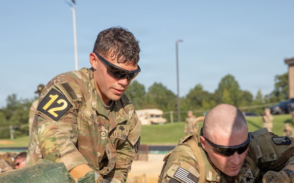 Two U.S. Army soldiers wearing OCP (Operational Camouflage Pattern) uniforms and numbered competition bibs (12 and 8) are working together to stack or arrange green sandbags. The activity appears to be part of a military competition or training exercise, likely a Best Warrior or similar skills competition. The setting is an outdoor military installation with other soldiers and buildings visible in the background.