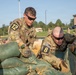 Two U.S. Army soldiers wearing OCP (Operational Camouflage Pattern) uniforms and numbered competition bibs (12 and 8) are working together to stack or arrange green sandbags. The activity appears to be part of a military competition or training exercise, likely a Best Warrior or similar skills competition. The setting is an outdoor military installation with other soldiers and buildings visible in the background.