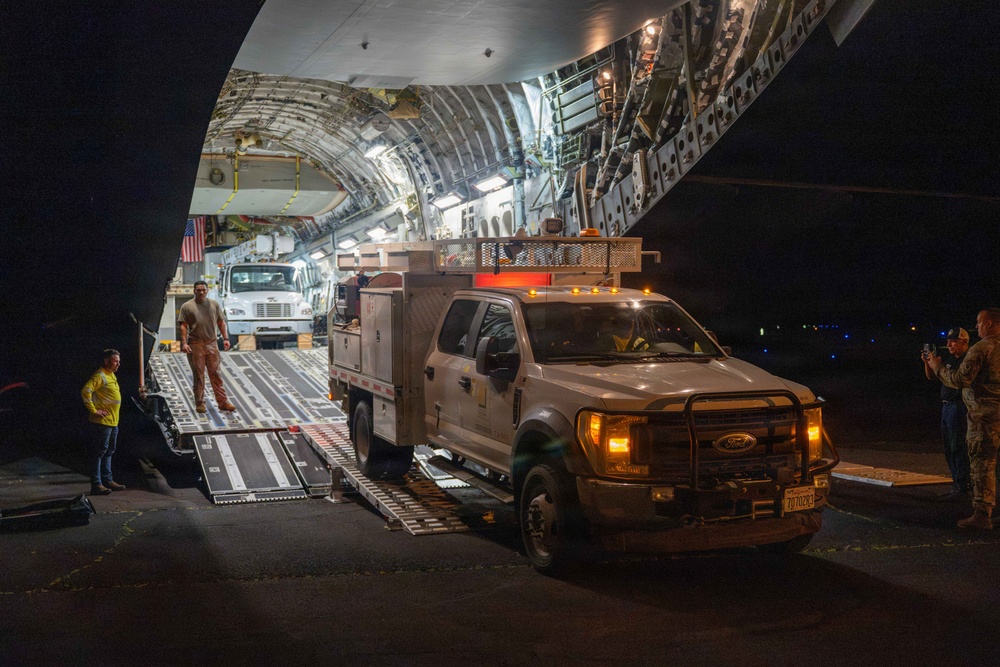 A nighttime military airlift operation showing vehicles being unloaded from a large military cargo aircraft, likely a C-17 Globemaster III or C-5 Galaxy. A white Ford F-450/550 utility/service truck with an aerial work platform is driving down the cargo ramp onto the tarmac. Additional vehicles, including a larger truck, are visible inside the illuminated cargo hold.