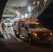 A nighttime military airlift operation showing vehicles being unloaded from a large military cargo aircraft, likely a C-17 Globemaster III or C-5 Galaxy. A white Ford F-450/550 utility/service truck with an aerial work platform is driving down the cargo ramp onto the tarmac. Additional vehicles, including a larger truck, are visible inside the illuminated cargo hold.