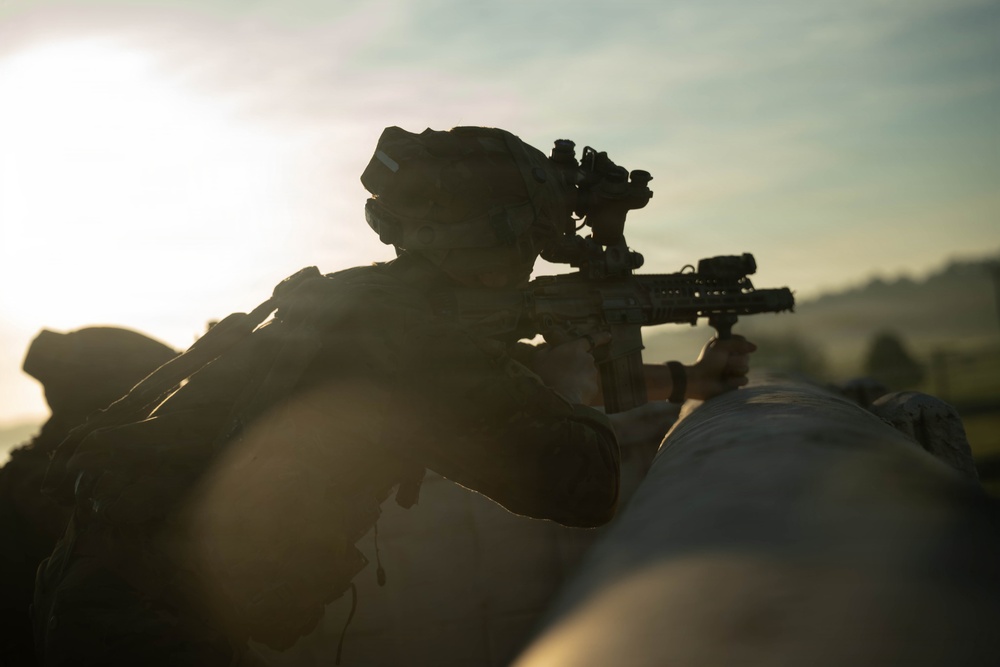 A heavily silhouetted soldier in full tactical gear is aiming a rifle equipped with optical sights and accessories over a barrier or surface. The image is taken during low-light conditions, likely at dawn or dusk, with strong backlighting creating a dramatic silhouette effect. A second figure is partially visible in the lower left of the frame.
