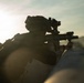 A heavily silhouetted soldier in full tactical gear is aiming a rifle equipped with optical sights and accessories over a barrier or surface. The image is taken during low-light conditions, likely at dawn or dusk, with strong backlighting creating a dramatic silhouette effect. A second figure is partially visible in the lower left of the frame.