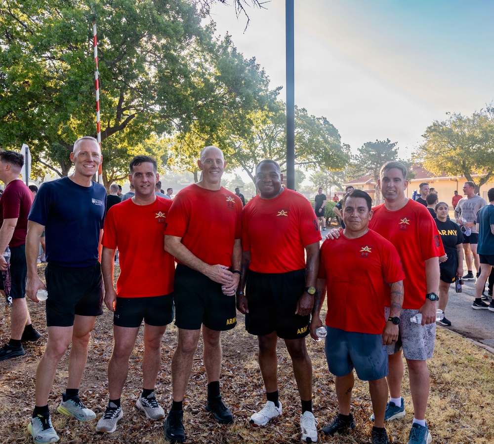 A group of six men pose together outdoors at what appears to be a military fitness event or run. Five of the men are wearing matching red t-shirts with a gold star insignia, consistent with U.S. Army unit PT (physical training) shirts, while one wears a navy blue shirt. The setting appears to be on a military installation with other participants visible in the background.