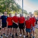 A group of six men pose together outdoors at what appears to be a military fitness event or run. Five of the men are wearing matching red t-shirts with a gold star insignia, consistent with U.S. Army unit PT (physical training) shirts, while one wears a navy blue shirt. The setting appears to be on a military installation with other participants visible in the background.