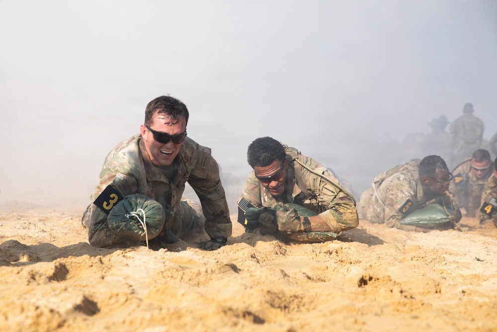 Multiple military personnel in camouflage uniforms are low-crawling through sandy terrain during what appears to be a training exercise or competition. Smoke or haze fills the background, creating a simulated combat environment. Participants are numbered with armbands, suggesting a structured competitive or evaluation event.