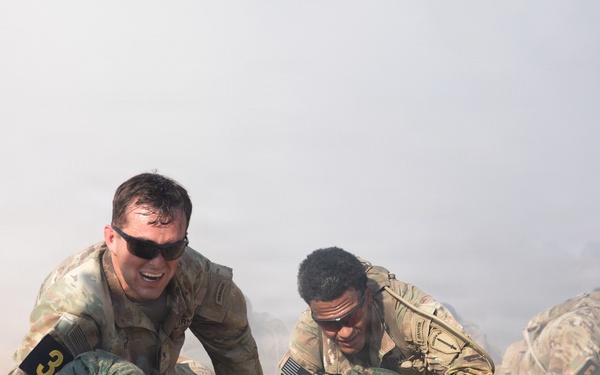 Multiple military personnel in camouflage uniforms are low-crawling through sandy terrain during what appears to be a training exercise or competition. Smoke or haze fills the background, creating a simulated combat environment. Participants are numbered with armbands, suggesting a structured competitive or evaluation event.