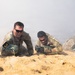Multiple military personnel in camouflage uniforms are low-crawling through sandy terrain during what appears to be a training exercise or competition. Smoke or haze fills the background, creating a simulated combat environment. Participants are numbered with armbands, suggesting a structured competitive or evaluation event.