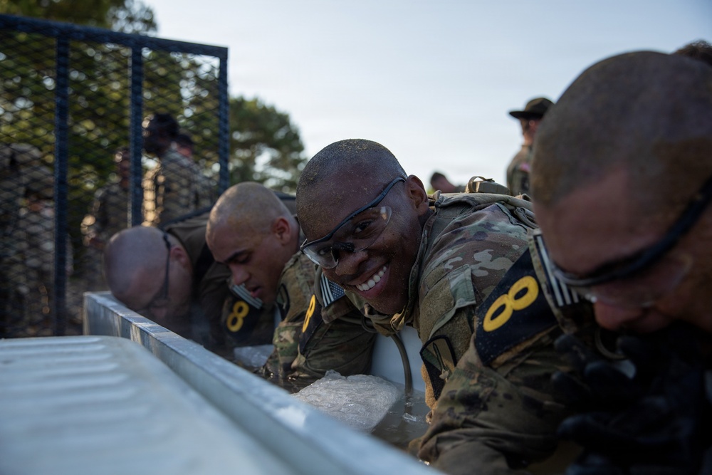 Military personnel in camouflage uniforms are participating in what appears to be a cold water immersion or ice bath endurance training exercise. The soldiers are wearing numbered bibs (visible '8' and '0' markings) and protective eyewear, suggesting a formal competition or assessment event. One soldier in the center is smiling broadly despite the apparent physical stress of the exercise.