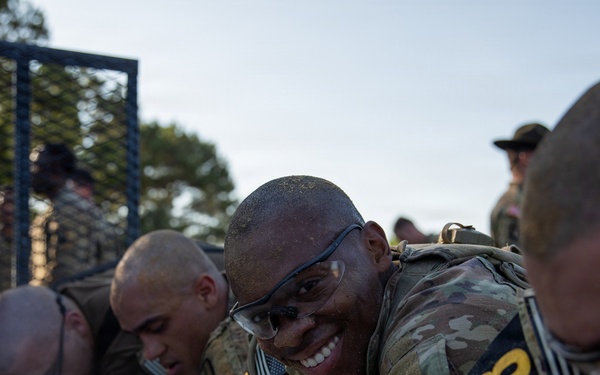 Military personnel in camouflage uniforms are participating in what appears to be a cold water immersion or ice bath endurance training exercise. The soldiers are wearing numbered bibs (visible '8' and '0' markings) and protective eyewear, suggesting a formal competition or assessment event. One soldier in the center is smiling broadly despite the apparent physical stress of the exercise.