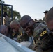 Military personnel in camouflage uniforms are participating in what appears to be a cold water immersion or ice bath endurance training exercise. The soldiers are wearing numbered bibs (visible '8' and '0' markings) and protective eyewear, suggesting a formal competition or assessment event. One soldier in the center is smiling broadly despite the apparent physical stress of the exercise.