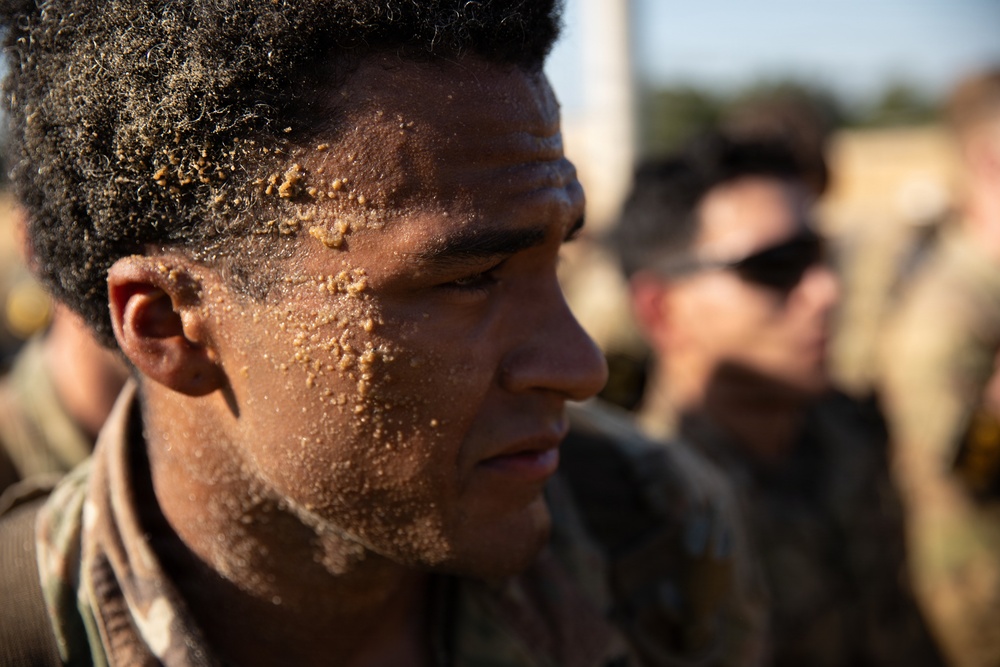 A close-up photograph of a military service member with sand covering his face and hair, suggesting recent physical training or a beach/surf exercise. He is wearing camouflage uniform and appears focused or fatigued. Other service members in camouflage are visible in the blurred background.